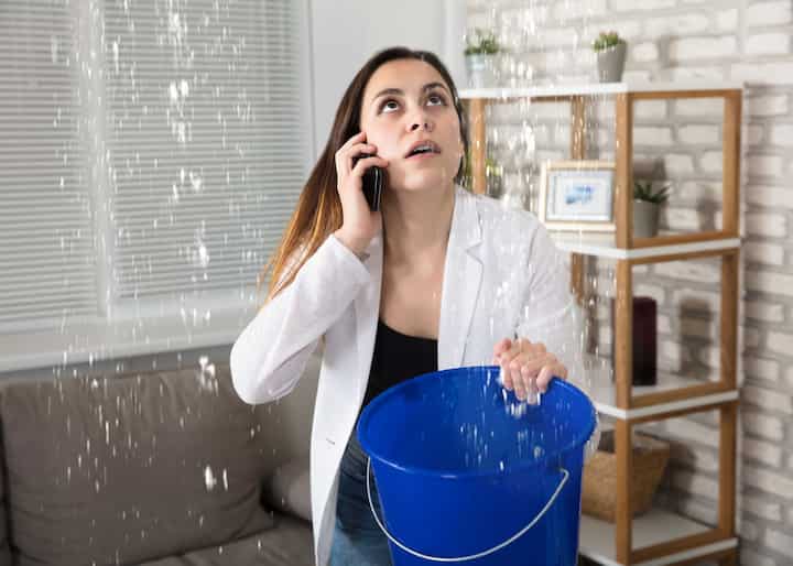 A woman calling water damage repair experts while holding a bucket trying to catch the water leaking from her ceiling in Augusta, GA